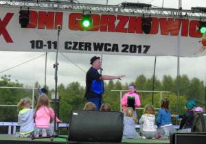 Meżczyzna w czarnym kapeluszu stoi na scenie i mówi przez mikrofon do dzieci