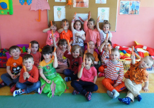 Dzieci siedzą na dywanie. W podniesionych rękach trzymjaą marchew.