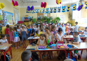 Dzieci siedzą w ławkach. Za nimi dzieci stoją.