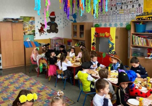 Dzieci w kolorowych strojach siedzą przy stolikach i jedzą poczęstunek.