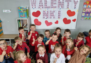Grupa dzieci pozuje do zdjęcia trzymając w rękach czerwone lizaki w kształcie serca. W tle na tablicy napis Walentynki.
