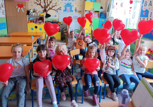 Grupa dzieci pozuje do zdjęcia siedząc na krzesełkach i trzymając w rękach w górze czerwone balony w kształcie serca.