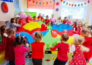 Grupa dzieci bawi się chustą an8imacyjną podrzucając na niej czerwony balon w kształcie serca.