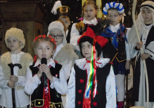 Dziewczynka i chłopiec są ubrani w ludowe stroje za nimi dzieci przebrane za anioły i pasterzy