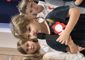 Na zdjęciu jest dziewczynka , która recytuje wiersz za nią stoi grupa dzieci biorąca udział w występie