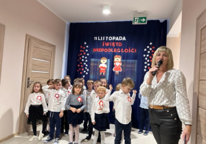 Na zdjęciu znajduje się Pani Dyrektor, która przemawia do publiczności oraz grupa dzieci biorąca udział w występie z okazji 11 Listopada