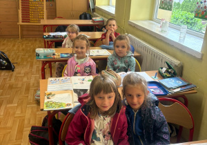 Przedszkolaki siedzący w szkolnych ławkach przy otwartych książkach.