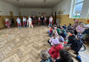 Grupa przedszkolaków oglądających występ uczniów na szkolnym korytarzu.