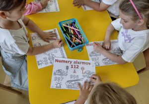 Przedszkolaki kolorujące przy stoliku obrazki dotyczące numerów alarmowych.