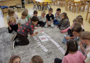 Przedszkolaki układające na dywanie karteczki dotyczące konieczności wybrania numeru alarmowego.