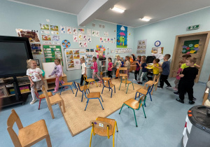 Dzieci w trakcie zabawy w sali, biegające pomiędzy rozstawionymi krzesełkami.