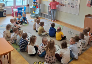Grupa przedszkolaków siedząca na dywanie wraz z nauczycielką omawiającą temat bezpieczeństwa w ruchu drogowym.