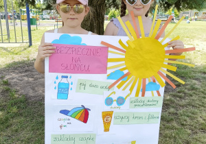 Dwie dziewczynki w nakryciach głowy i okularach przeciwsłonecznych, trzymające plakat "Bezpiecznie na słońcu".