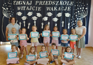 Grupa dzieci trzymających dyplomy wraz z dwiema nauczycielkami na tle dekoracji z okazji zakończenia roku.