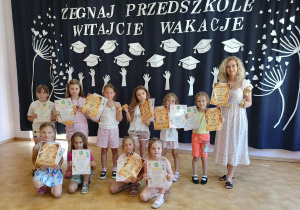 Dziewczynki z grupy tanecznej prezentujące dyplomy i medale wraz z opiekunką trzymającą puchar na tle dekoracji z okazji zakończenia roku.